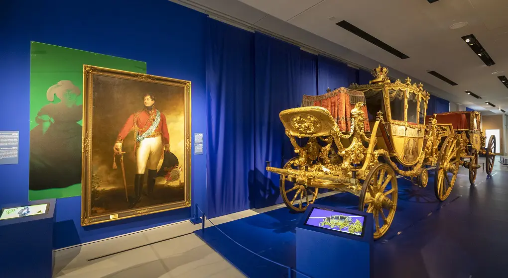 The photo shows an exhibition room in the Museum Schloss Herrenhausen in Hanover. In the foreground stands an ornate, golden state carriage with intricate details. Next to it hangs a large portrait of George IV wearing a red uniform. The deep blue walls enhance the regal atmosphere of the display.