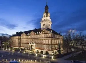 La photo montre un grand château. Le château a beaucoup de fenêtres et une haute tour. Le soir, les lumières sont jolies. Le château se trouve à Wolfenbüttel. Autrefois, des ducs vivaient ici. Aujourd’hui, on peut visiter le château.