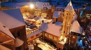 Das Bild zeigt den Weihnachtsmarkt in Braunschweig. Alles ist mit Schnee bedeckt. Viele kleine Stände stehen auf dem Platz. Lichterketten leuchten warm im Dunkeln. Menschen spazieren zwischen den Ständen. Ein großes Gebäude mit Turm steht am Rand. Die Stimmung ist winterlich und festlich.