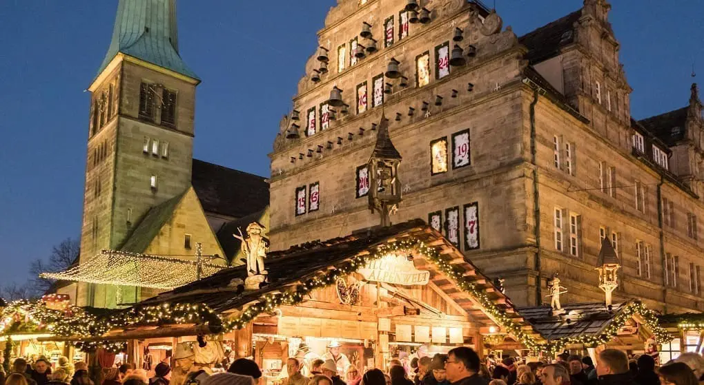 Auf dem Bild sieht man einen Weihnachtsmarkt in Hameln. Es ist schon dunkel, aber viele Lichter leuchten. Man sieht viele Menschen und schöne Buden. Ein großes, altes Haus ist im Hintergrund. Es ist eine festliche Stimmung.