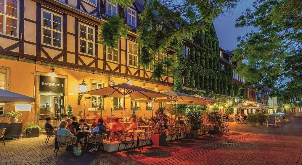 Das Bild zeigt den Ballhof in Hannover am Abend. Die Fachwerkhäuser sind beleuchtet. Viele Menschen sitzen draußen vor einem Restaurant. Über den Tischen sind große Schirme. Bäume und Pflanzen sind rundherum.