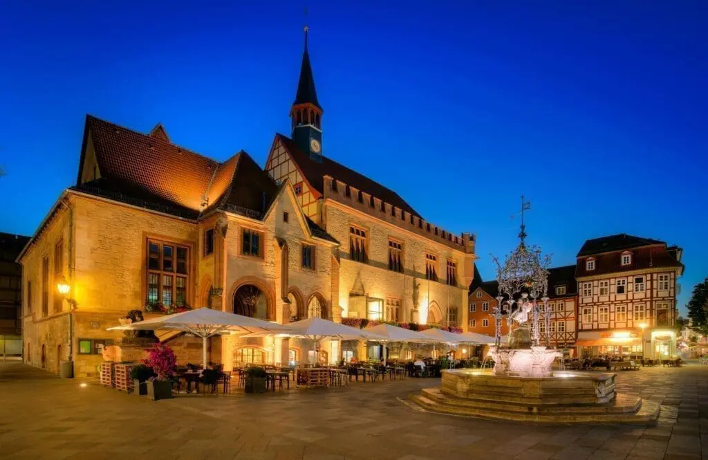 The picture shows the Old Town Hall in Göttingen. The large building is made of stone. It has many windows and a red roof. Next to the town hall is a beautiful fountain. In the evening, many lights shine. The town hall is a famous landmark in the city.