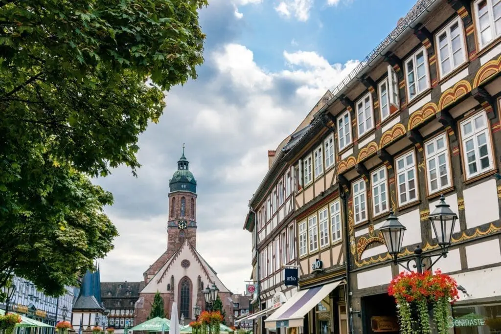 Auf dem Bild sieht man den Marktplatz von Einbeck. In der Mitte steht eine große Kirche mit einem Turm. Rundherum sind alte Fachwerkhäuser mit schönen Verzierungen. Es gibt auch viele Blumen und Sonnenschirme auf dem Platz.