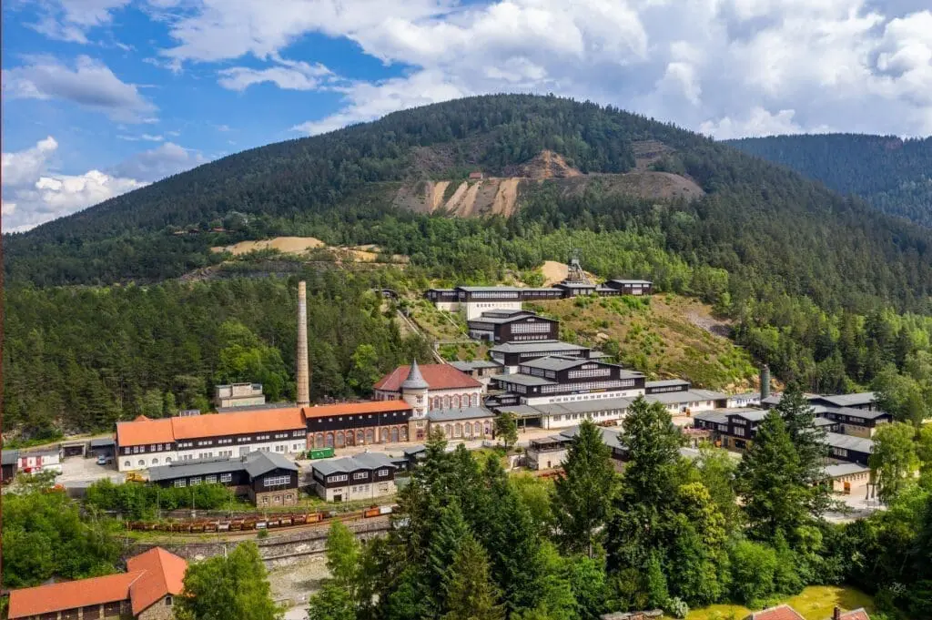 En la imagen se ven muchos edificios en una montaña. Son parte de una mina antigua. Antes se sacaban metales de esta montaña. La montaña tiene muchos árboles. Se ve una chimenea alta. Hace buen tiempo con sol y algunas nubes. Es la mina de Rammelsberg en Goslar.