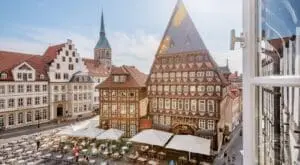 L’image montre la place du marché à Hildesheim. On voit de belles maisons anciennes. Au centre se trouve le grand Knochenhaueramtshaus. C’est une maison à colombages avec beaucoup de fenêtres. À gauche, il y a la maison blanche Rolandstube. En arrière-plan, une église avec une haute tour. Il fait clair et calme.