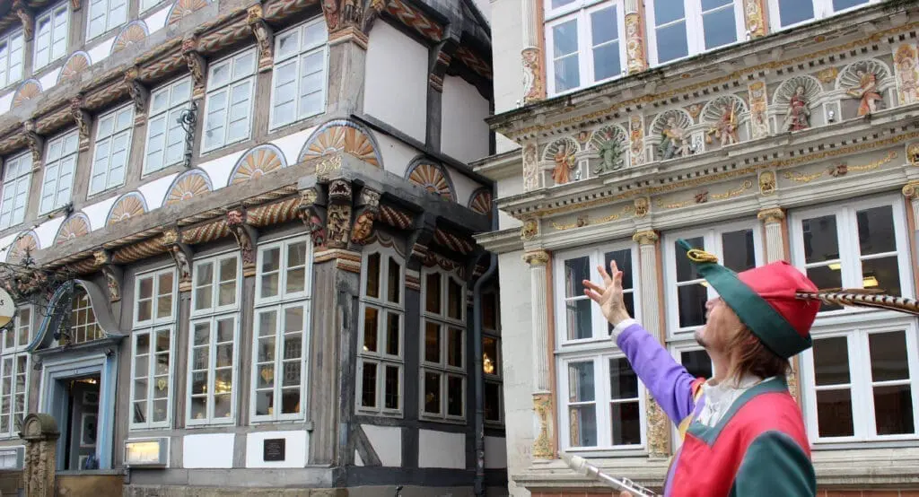 Billedet viser en byvandring i Hameln. En mand har et farverigt kostume på. Han ligner rottefængeren fra den kendte historie. Han peger på et gammelt bindingsværkshus. Husene har mange flotte detaljer.