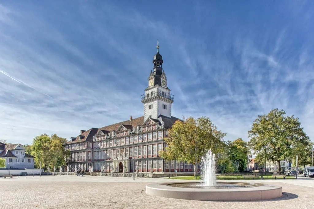 Das Bild zeigt das Schloss in Wolfenbüttel. Das Schloss ist sehr groß und hat viele Fenster. Ein hoher Turm steht in der Mitte. Vorne sind Bäume mit grünen und gelben Blättern. Der Himmel ist blau und es ist ein schöner Tag.