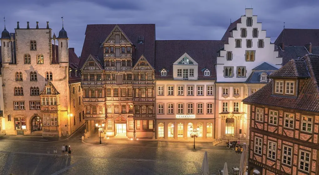Das Bild zeigt den Marktplatz in Hildesheim. Die Häuser sind schön beleuchtet. Man sieht viele alte Gebäude. Es ist schon Abend.