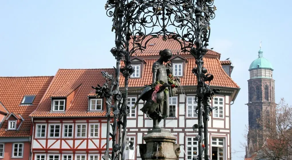 Das Bild zeigt die Gänseliesel-Figur in Göttingen. Die Figur steht auf einem Brunnen. Im Hintergrund sieht man Fachwerkhäuser und einen Kirchturm.