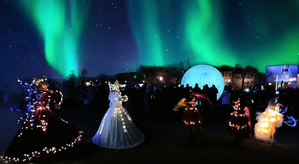 The picture shows a dance show in Celle. The show is in the evening. The dancers wear glowing dresses. Green northern lights are in the sky. Many people watch the show. It is a special experience.