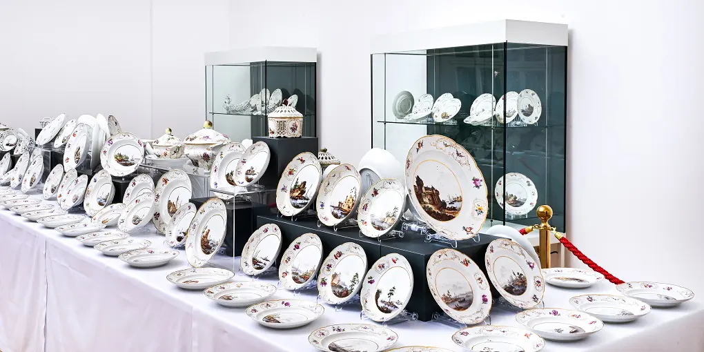 Bilden visar konstnärligt målade porslinstallrikar. Tallrikarna står på ett vitt bord. Det finns färgglada bilder på tallrikarna. Tallrikarna är en del av en utställning på residensmuseet i Wolfenbüttel.