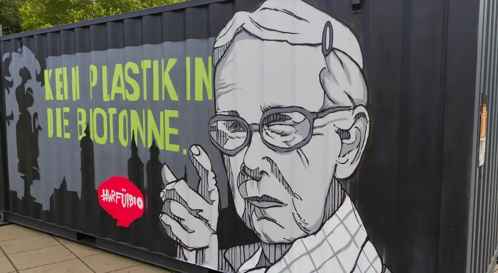 Bilden visar graffiti på en stor container. En äldre kvinna med glasögon pekar med fingret. Bredvid står det: “Kein Plastik in die Biotonne.” Det finns också en röd cirkel med #ARFUIBIO. I bakgrunden syns mörka byggnader. Budskapet är: Plast ska inte läggas i matavfallet.