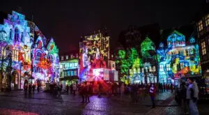 The picture shows a city at night. Colorful lights and images are projected onto the old houses. Many people stand on the square and watch. The atmosphere is bright and festive.