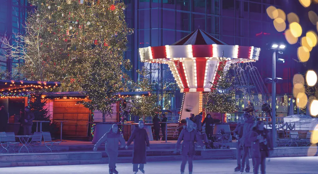 The picture shows a winter scene in the Autostadt. There is a big Christmas tree with many lights. Next to it is a carousel with red and white stripes. Many lights decorate the place. In the front, people are ice skating.