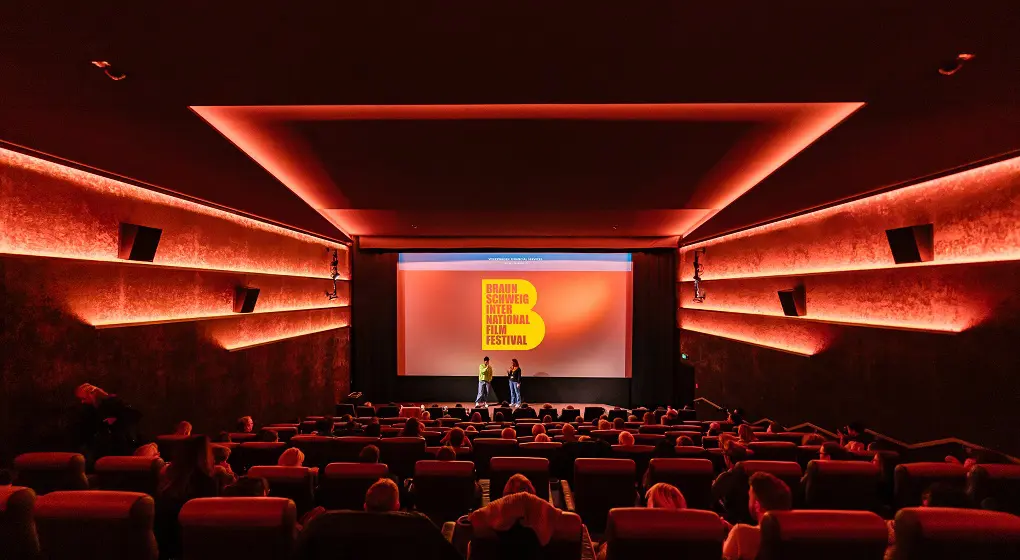 Salle de cinéma avec éclairage mural rouge, public, scène et écran lors du Braunschweig International Film Festival.