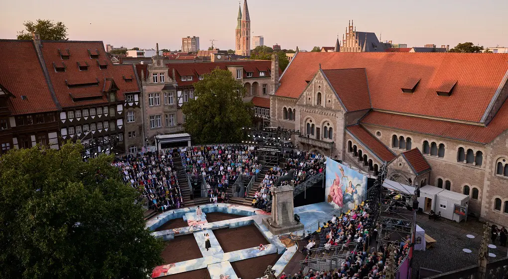 Muchas personas están sentadas en un teatro al aire libre en Brunswick. En el escenario actúan actores. Al fondo se ven casas antiguas y una iglesia.
