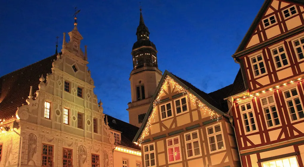 Auf dem Bild sieht man alte Häuser am Abend. Die Fachwerkhäuser und ein Barockhaus sind festlich mit Lichtern geschmückt. Im Hintergrund steht ein hoher Kirchturm vor dem dunklen, blauen Himmel. Das Bild wirkt ruhig und weihnachtlich.