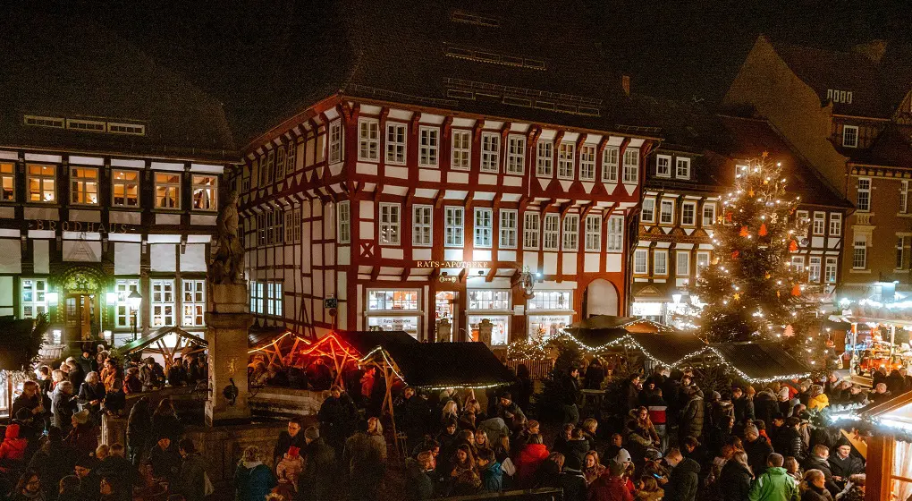 Auf dem Bild sieht man einen Weihnachtsmarkt. Viele Menschen stehen vor kleinen Holzbuden. Es gibt ein großes, beleuchtetes Weihnachts­baum. Die Häuser im Hintergrund sind alt und schön. Alles ist hell geschmückt und festlich.