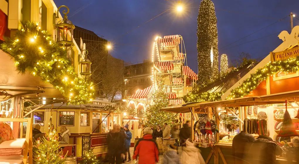 En julmarknad på kvällen med stånd, julgranar, ljus och ett pariserhjul.