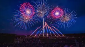 Ein Feuerwerk am Abendhimmel: Mehrere bunte Explosionen in Rot und Blau leuchten über einer Menschenmenge in einer Gartenanlage.