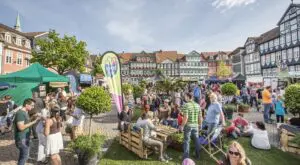 Viele Menschen feiern auf einem Platz mit alten Fachwerkhäusern. Es gibt bunte Stände mit Essen und Getränken. Menschen sitzen, essen, trinken und genießen das Fest.