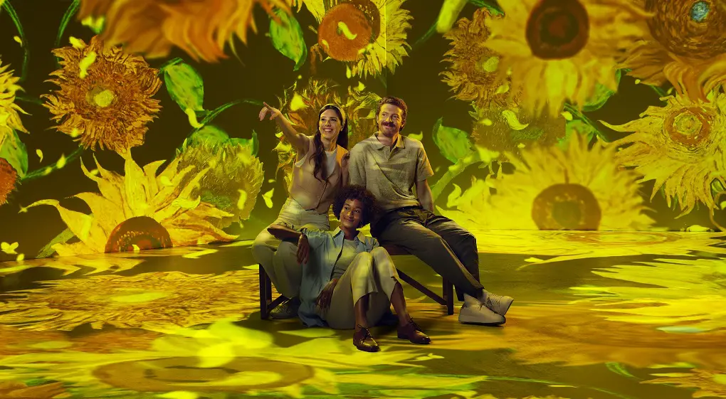 Tres personas están sentadas y sonrientes en una sala completamente rodeada por motivos de girasoles de Van Gogh en tonos amarillos brillantes. Las proyecciones cubren paredes y suelo, creando una experiencia artística inmersiva.