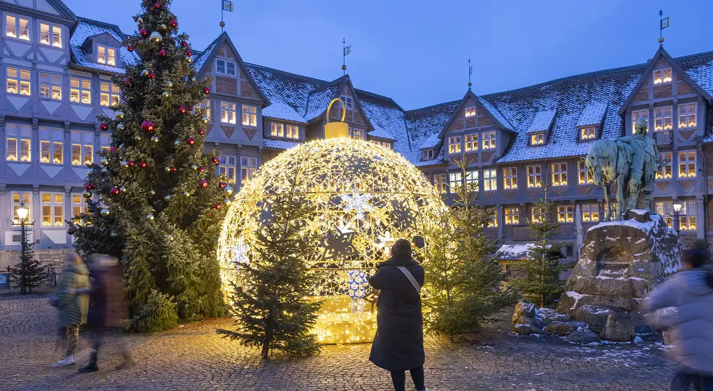 Auf dem Stadtmarkt in Wolfenbüttel steht ein großes Lichtobjekt in Form einer Weihnachtskugel. Daneben steht ein Weihnachtsbaum. Im Hintergrund sind Fachwerkhäuser mit vielen Lichtern in den Fenstern. Menschen laufen über den Platz.