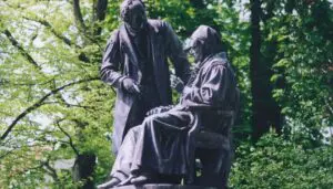 Das Gauß-Weber-Denkmal in Göttingen zeigt die Bronzefiguren der Wissenschaftler Carl Friedrich Gauß und Wilhelm Weber. Gauß sitzt auf einem Stuhl, während Weber neben ihm steht. Im Hintergrund sind grüne Bäume und Sträucher zu sehen.