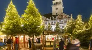 Mercado navideño de Wolfenbüttel al anochecer, con abetos iluminados, puestos de madera y visitantes frente a un castillo histórico.