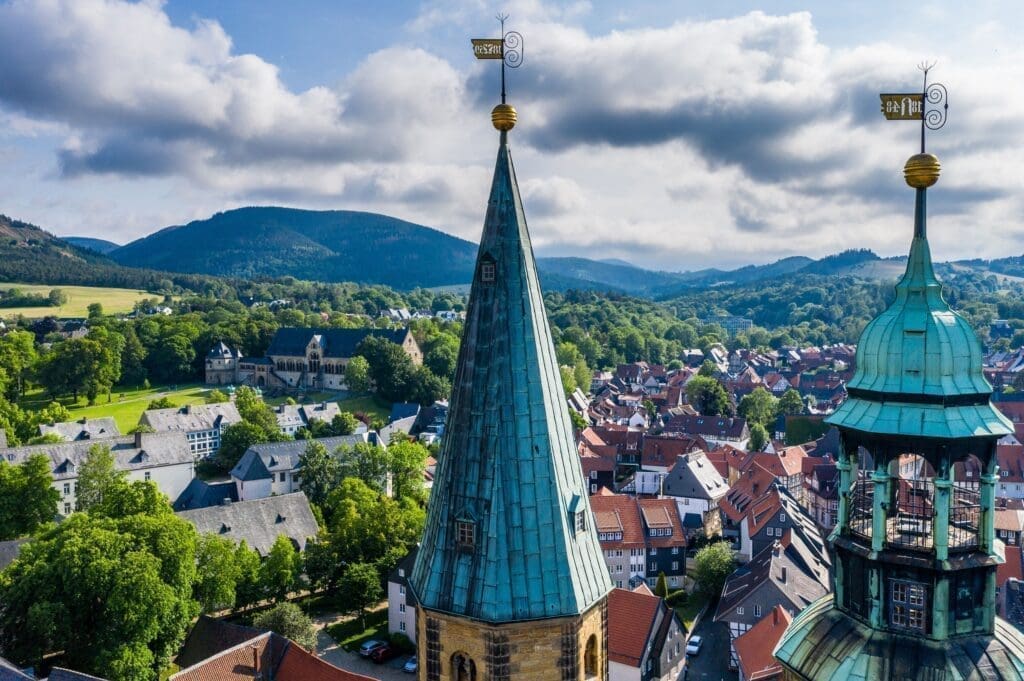 Das Bild zeigt die Stadt Goslar von oben. Man sieht viele rote Dächer und grüne Bäume. Vorne sind zwei hohe Kirchtürme mit grünen Dächern. Im Hintergrund sieht man Berge und Wälder. Es ist bewölkt, aber hell. Das Bild wirkt ruhig und schön.