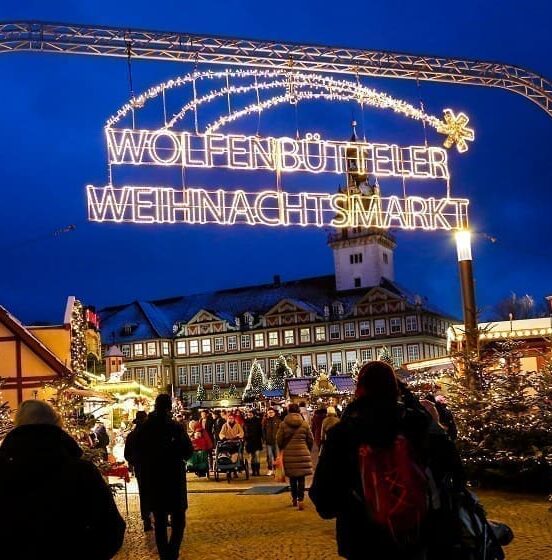 La imagen muestra el mercado navideño en Wolfenbüttel. Muchas personas caminan por el mercado. Hay muchas luces y árboles de Navidad. El ambiente es festivo. En el fondo se ve el castillo.